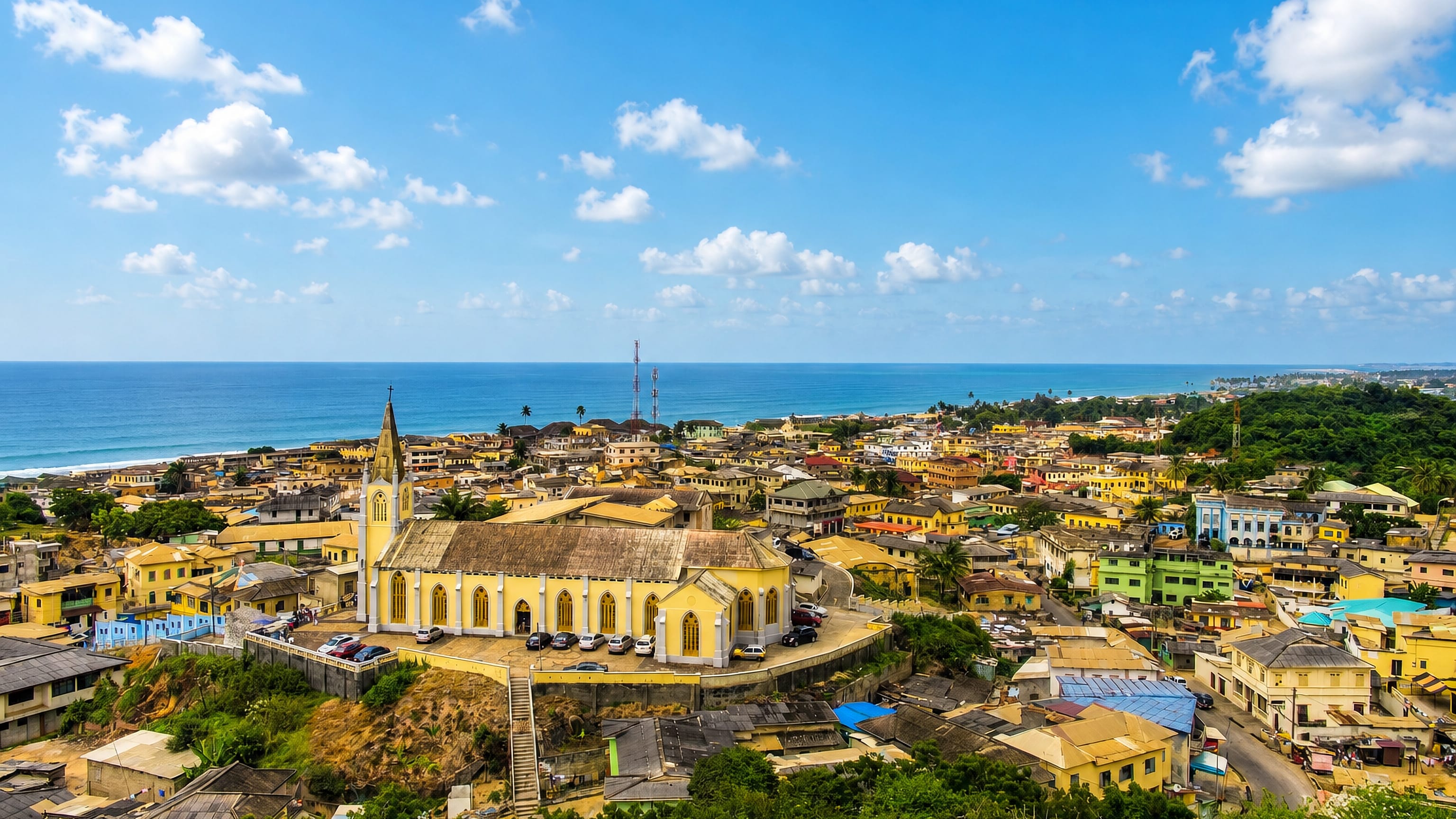 Hilltop church above the colorful low-rise rooftops of Cape Coast, Ghana, with the Atlantic beyond.