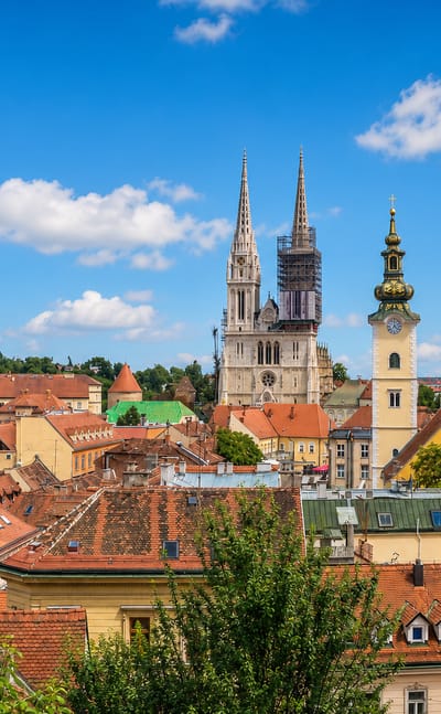 Zagreb's twin neo-Gothic cathedral spires rising above the old town's red-tiled roofs and a church tower under a blue sky.