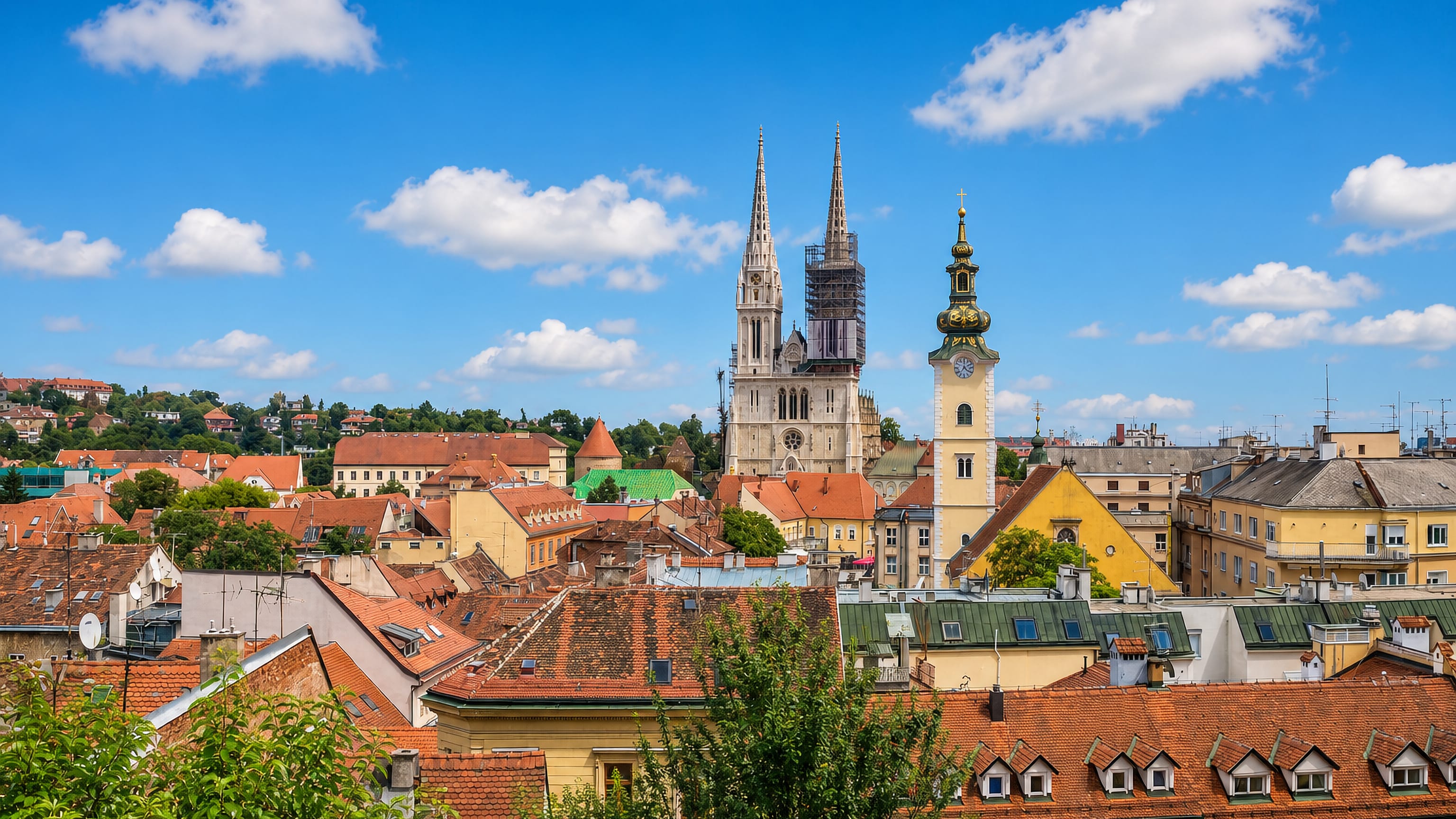 Zagreb's twin neo-Gothic cathedral spires rising above the old town's red-tiled roofs and a church tower under a blue sky.
