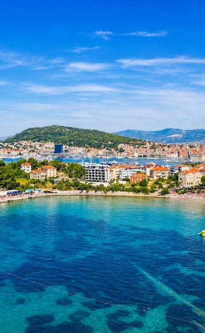 Aerial of Split's Bacvice cove with shallow turquoise water and sandy beach backed by the city peninsula and harbor ferry.