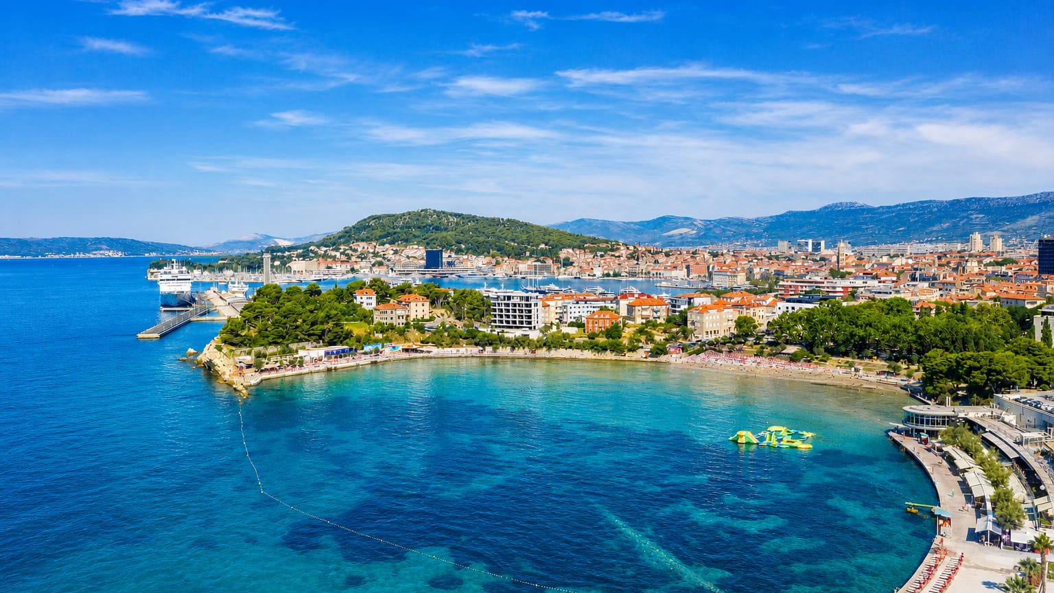 Aerial of Split's Bacvice cove with shallow turquoise water and sandy beach backed by the city peninsula and harbor ferry.