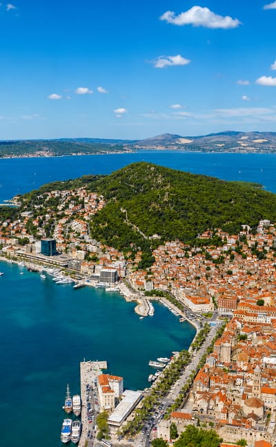 Aerial view of Split's peninsula, red-roofed old town and marina beside the turquoise Adriatic with wooded Marjan hill.