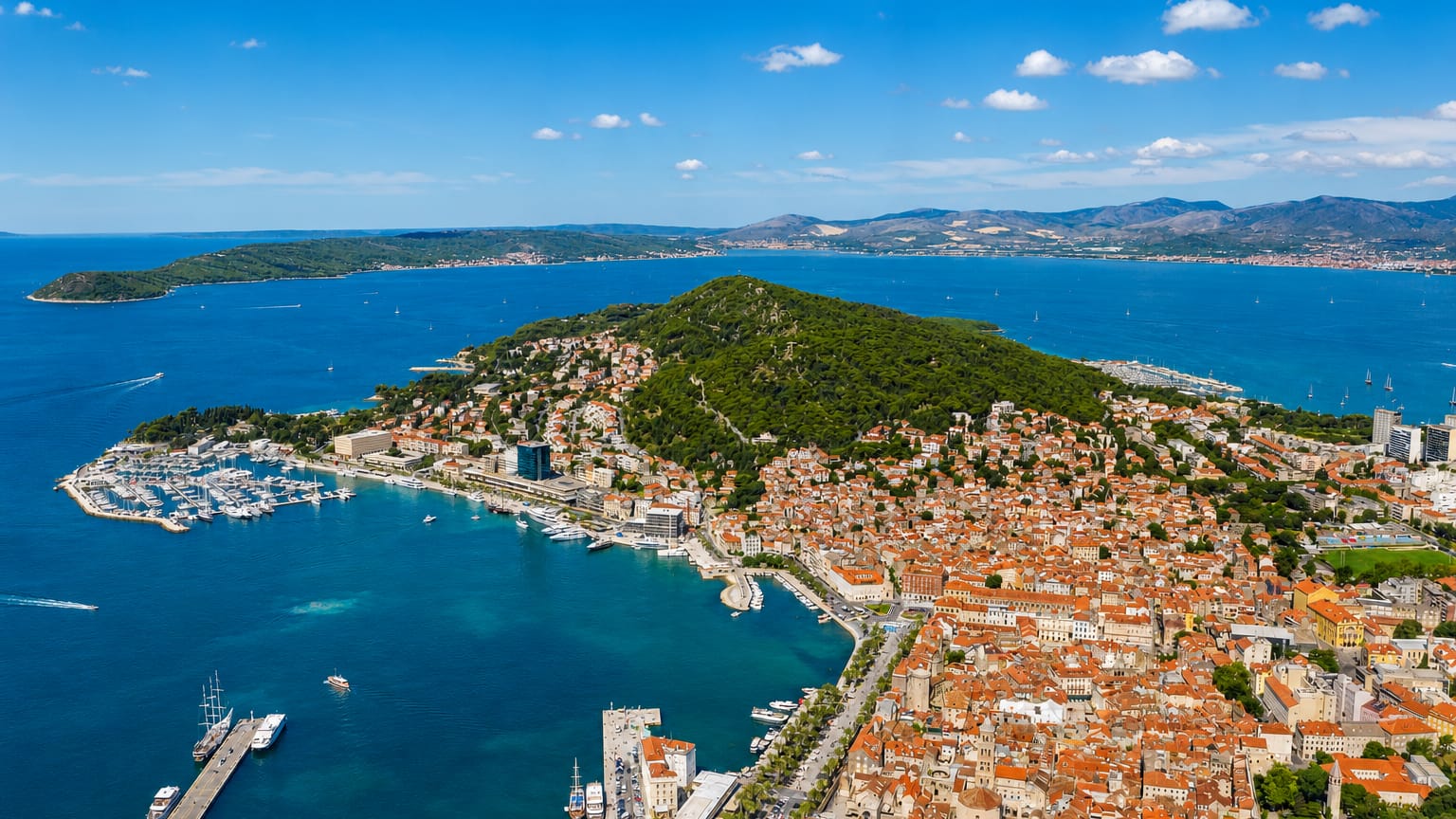 Aerial view of Split's peninsula, red-roofed old town and marina beside the turquoise Adriatic with wooded Marjan hill.