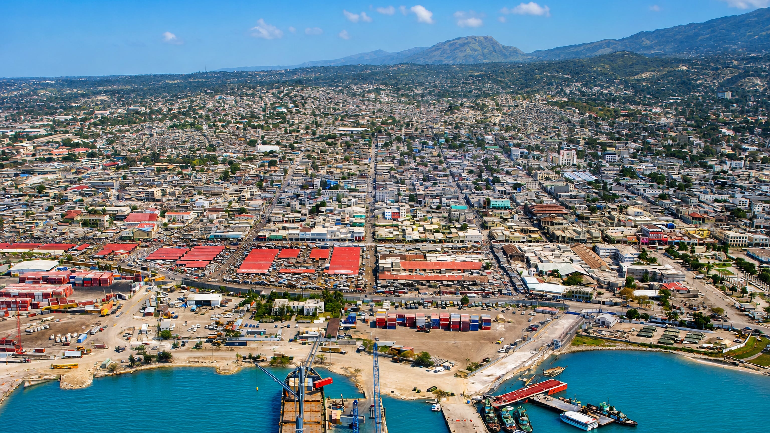 Aerial view of Port-au-Prince's container port and red-roofed market sheds, with the city rising to green mountains.