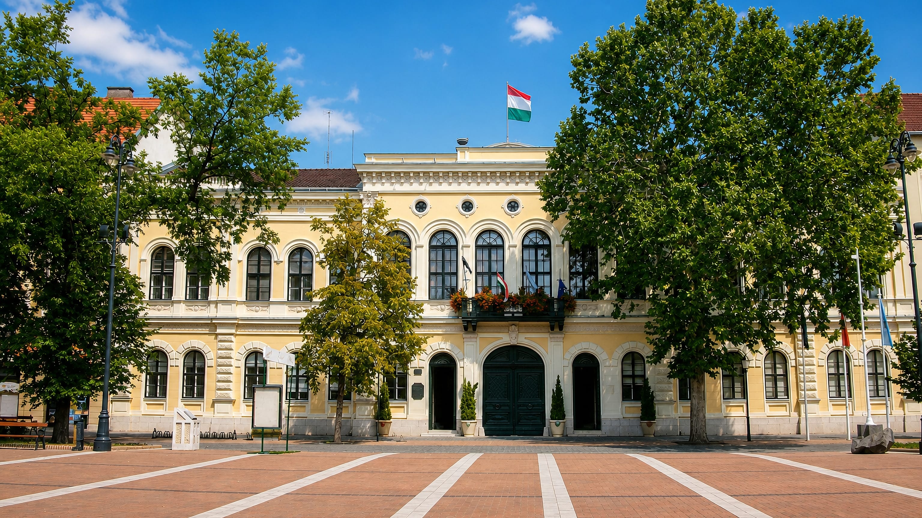 The yellow neoclassical town hall of Békéscsaba, Hungary, flying the national flag over its paved main square.