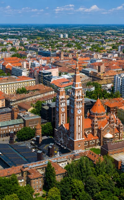Aerial view of Szeged's twin-towered brick Votive Church and Dom Square amid red-roofed streets.