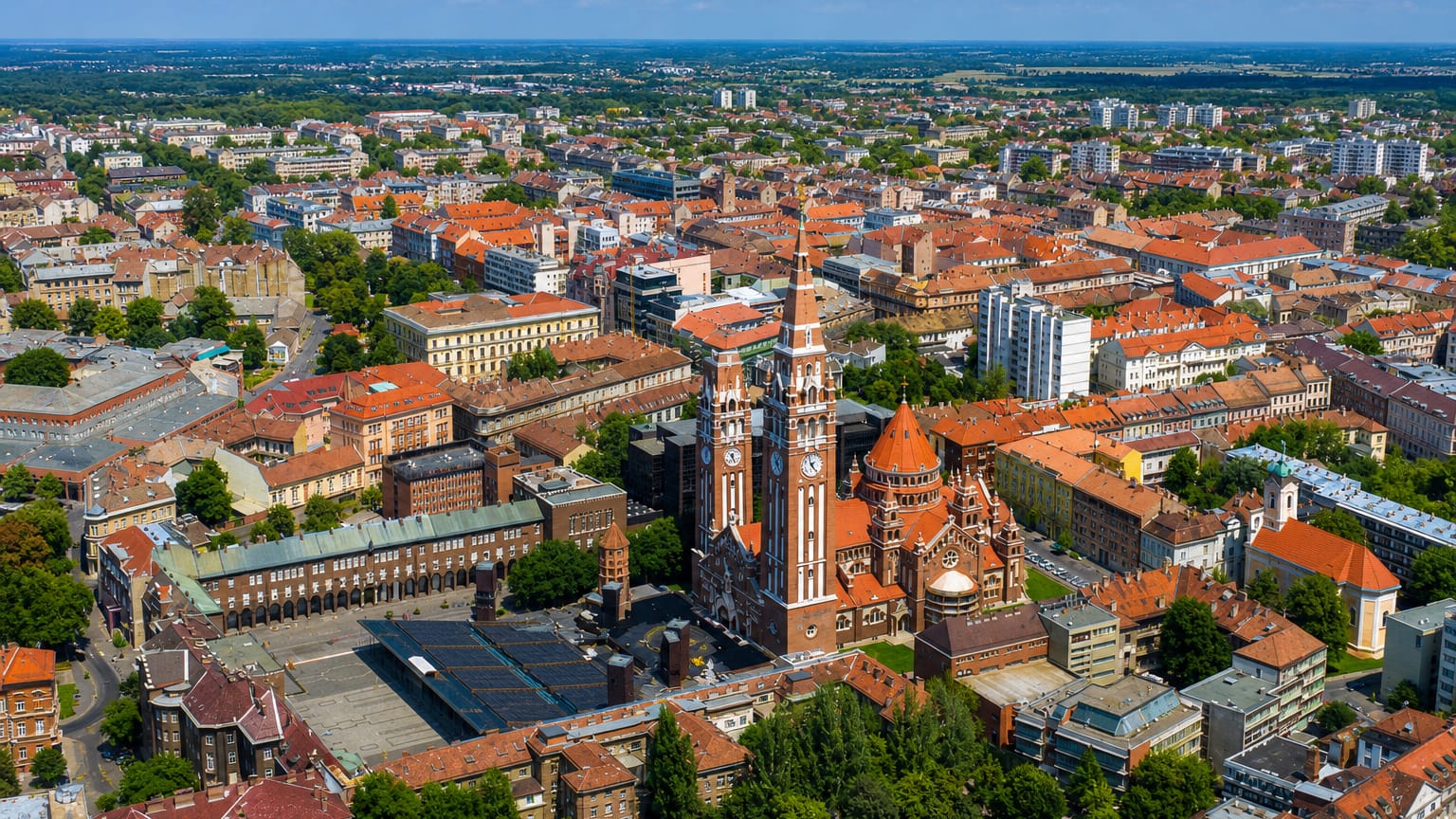 Aerial view of Szeged's twin-towered brick Votive Church and Dom Square amid red-roofed streets.
