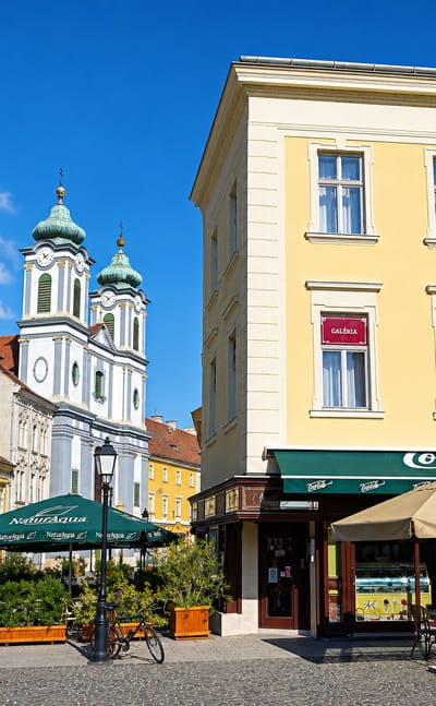 Szekesfehervar old-town corner with baroque twin church towers and a cafe terrace on a cobbled square.