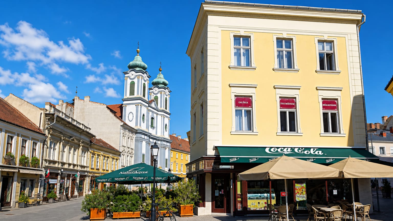 Szekesfehervar old-town corner with baroque twin church towers and a cafe terrace on a cobbled square.