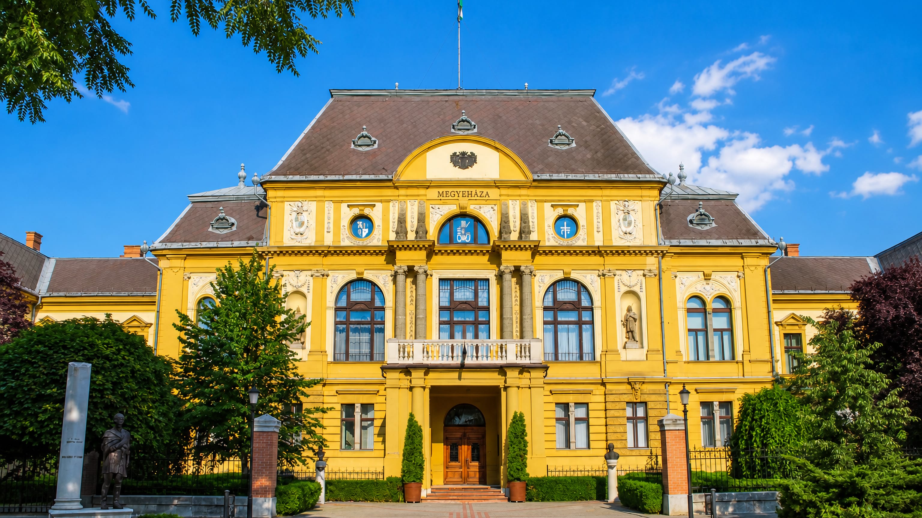 The yellow Megyehaza county hall in Nyiregyhaza, Hungary, fronted by clipped gardens and statues under blue sky.