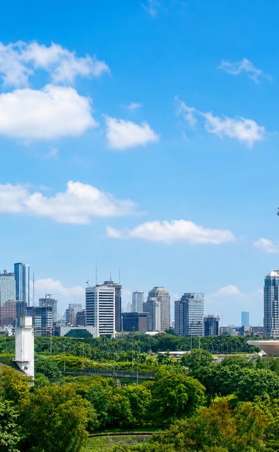 Jakarta skyline with the National Monument (Monas) tower rising above CBD high-rises and green parkland under a bright sky.