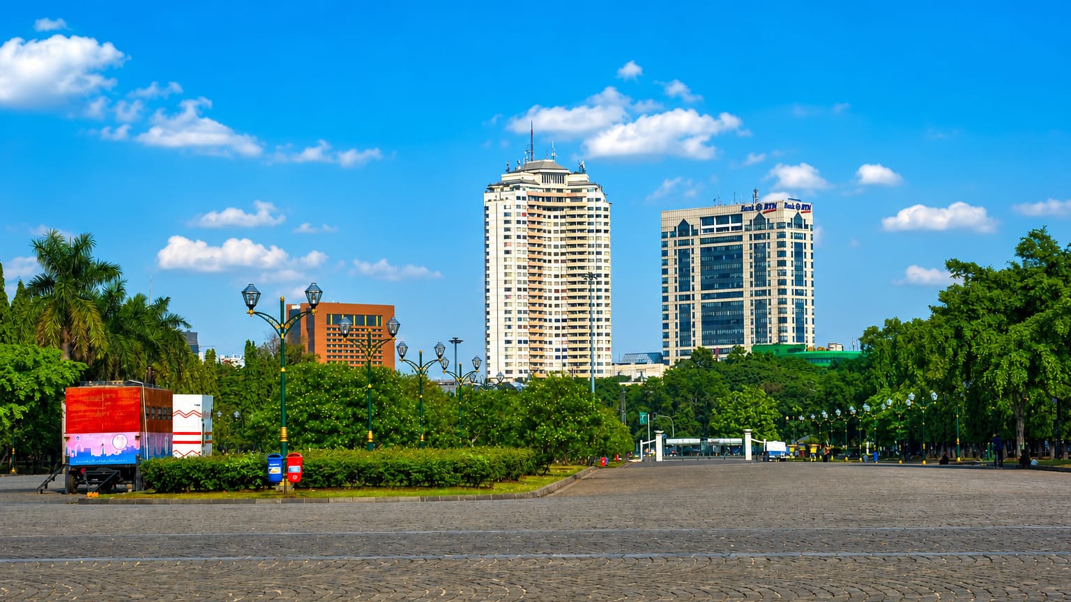 A broad paved plaza fringed by trees fronts office towers in Central Jakarta under a bright blue sky.
