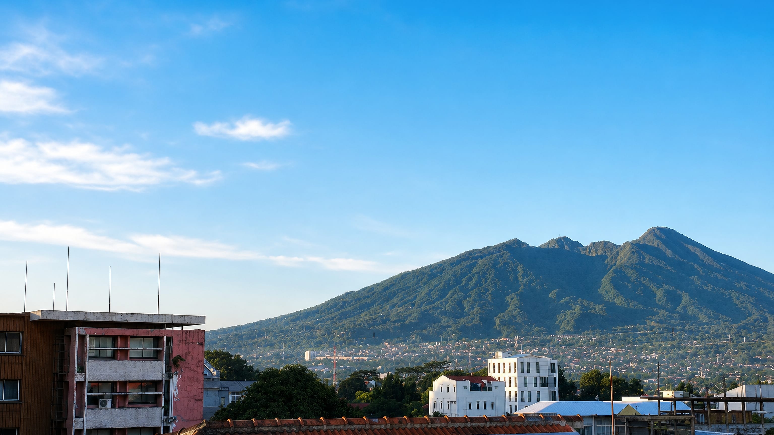 Rooftops of Bogor, West Java, below the forested twin peaks of Mount Salak under a clear sky.