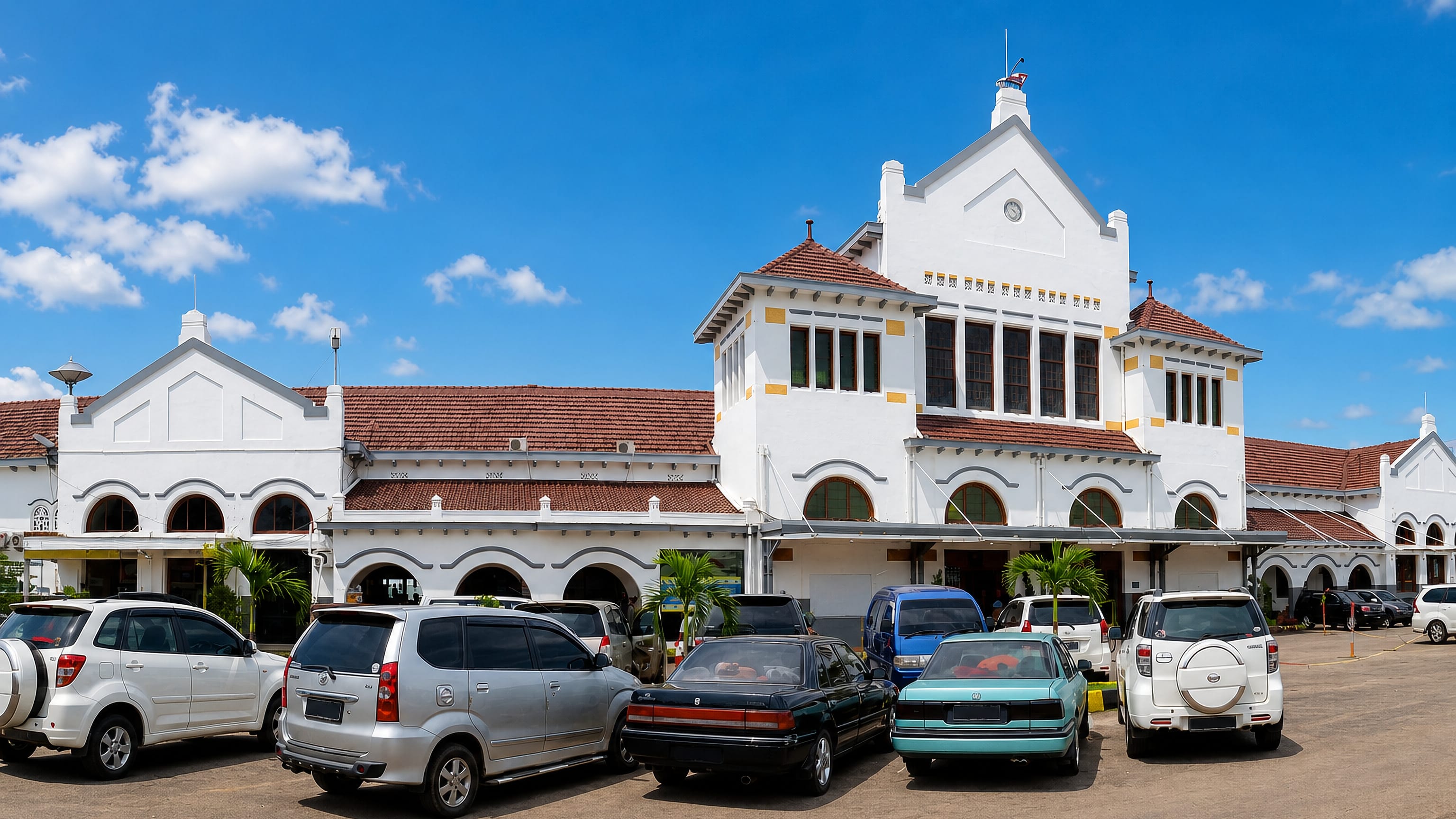 The white colonial facade and clock gable of Cirebon's Kejaksan railway station above a full car park.