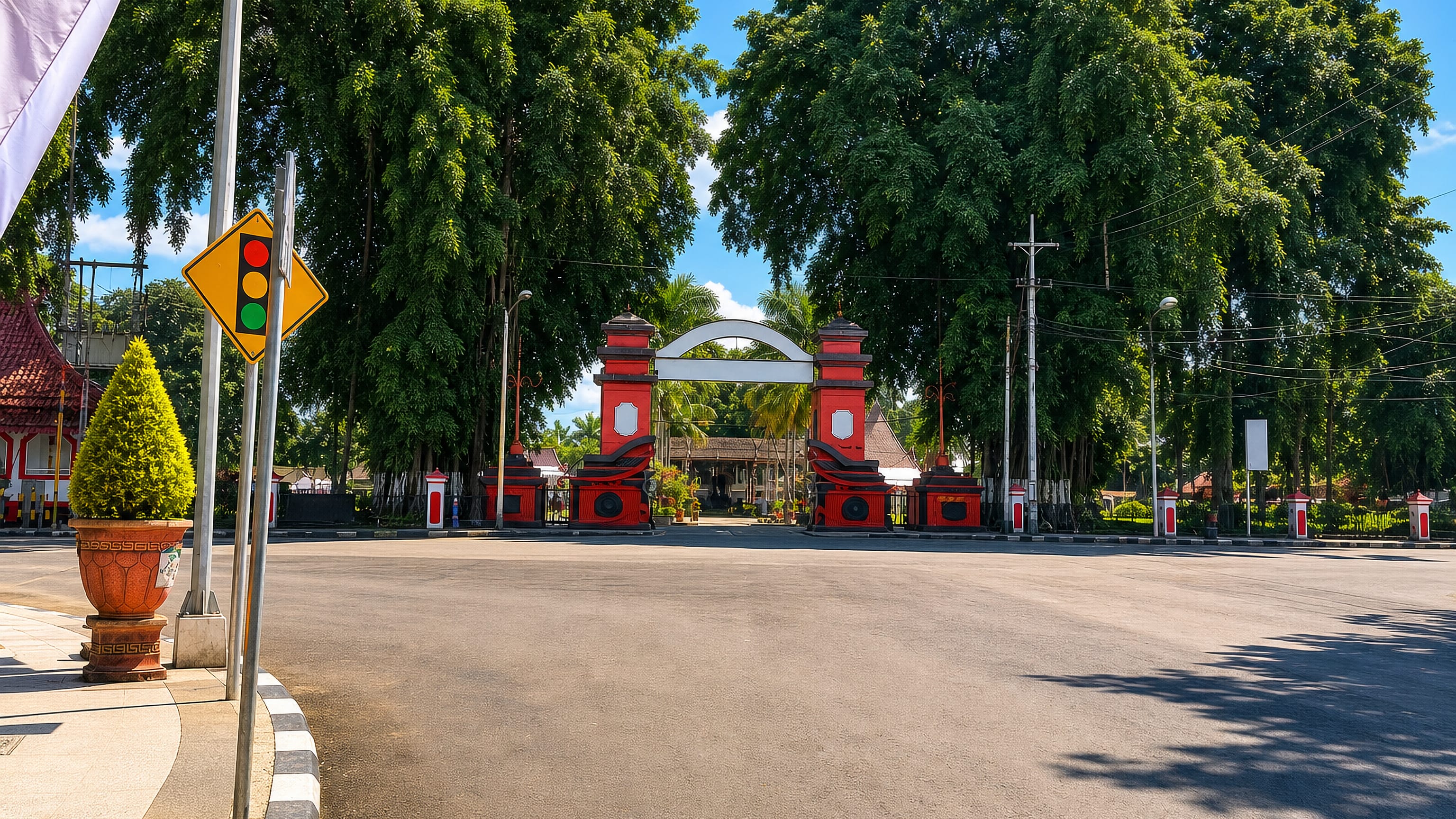 Red-and-white ceremonial gateway framed by tall shade trees on a quiet street in Blitar, East Java.