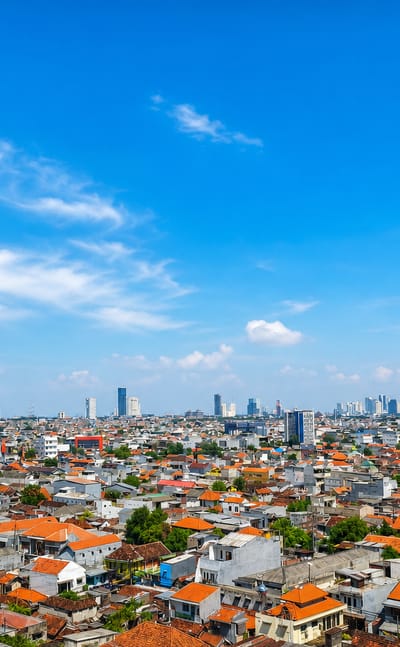 Surabaya cityscape of dense red-roofed houses stretching to distant high-rise towers under a bright blue sky.