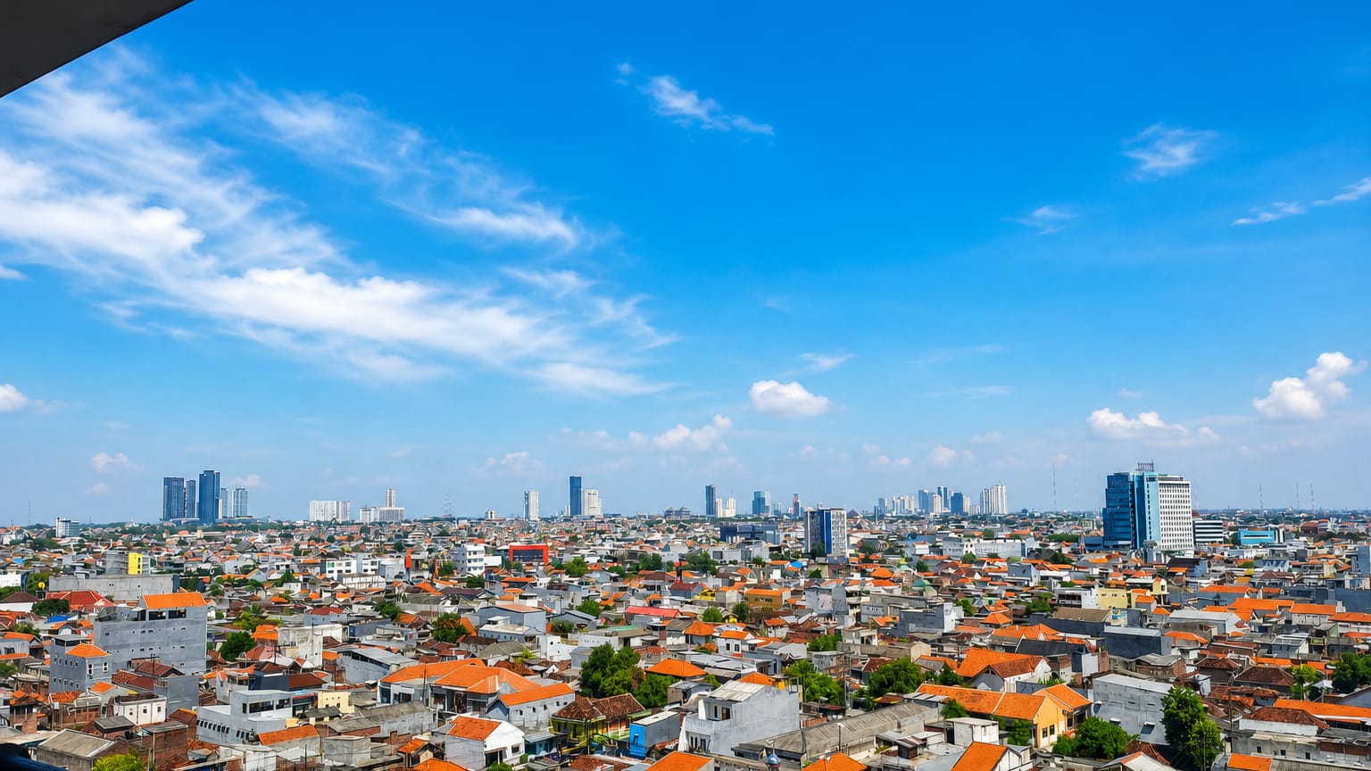 Surabaya cityscape of dense red-roofed houses stretching to distant high-rise towers under a bright blue sky.