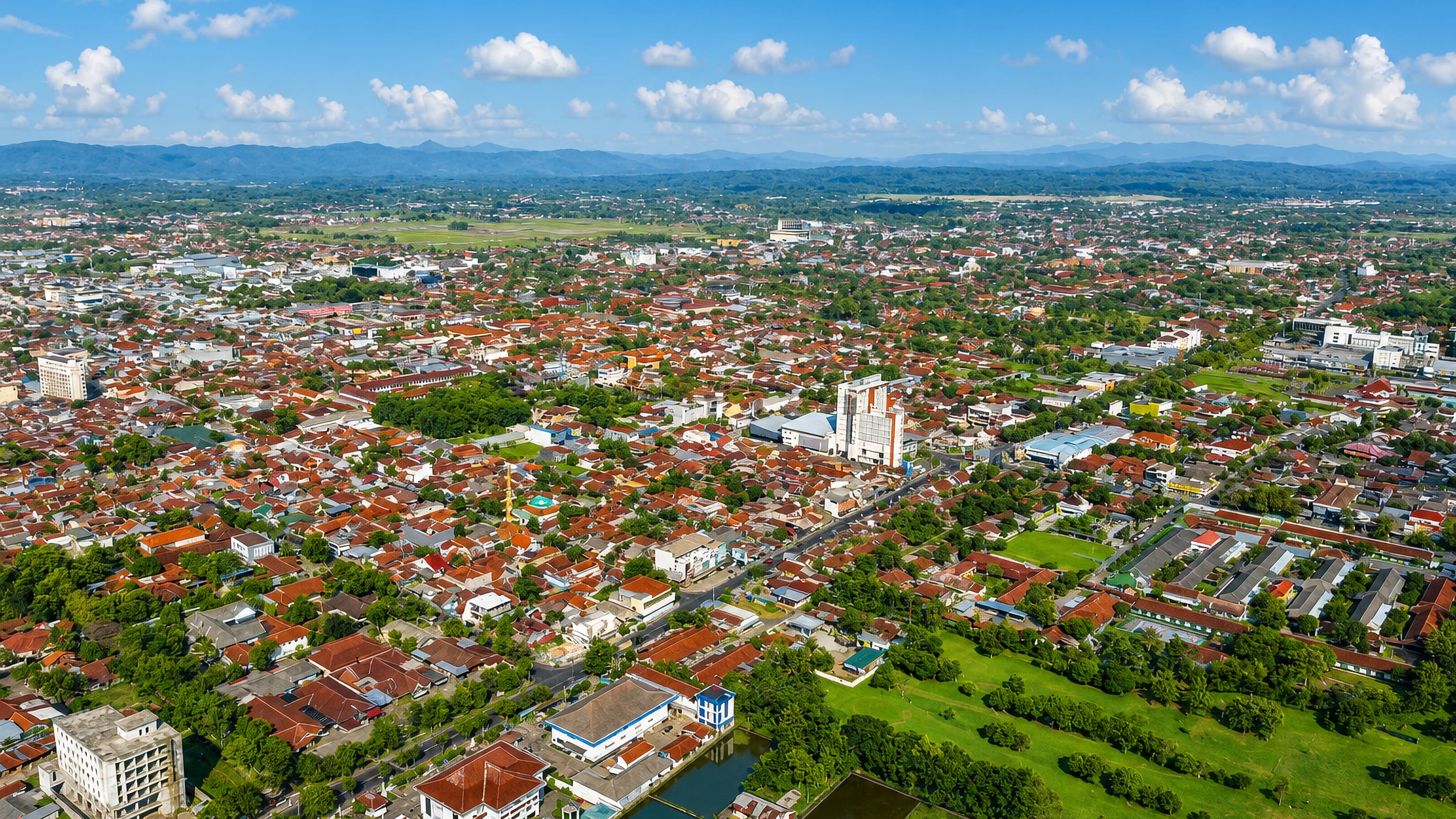 Aerial view of Purwokerto's red-tiled rooftops and green fields with the hills of Central Java behind.