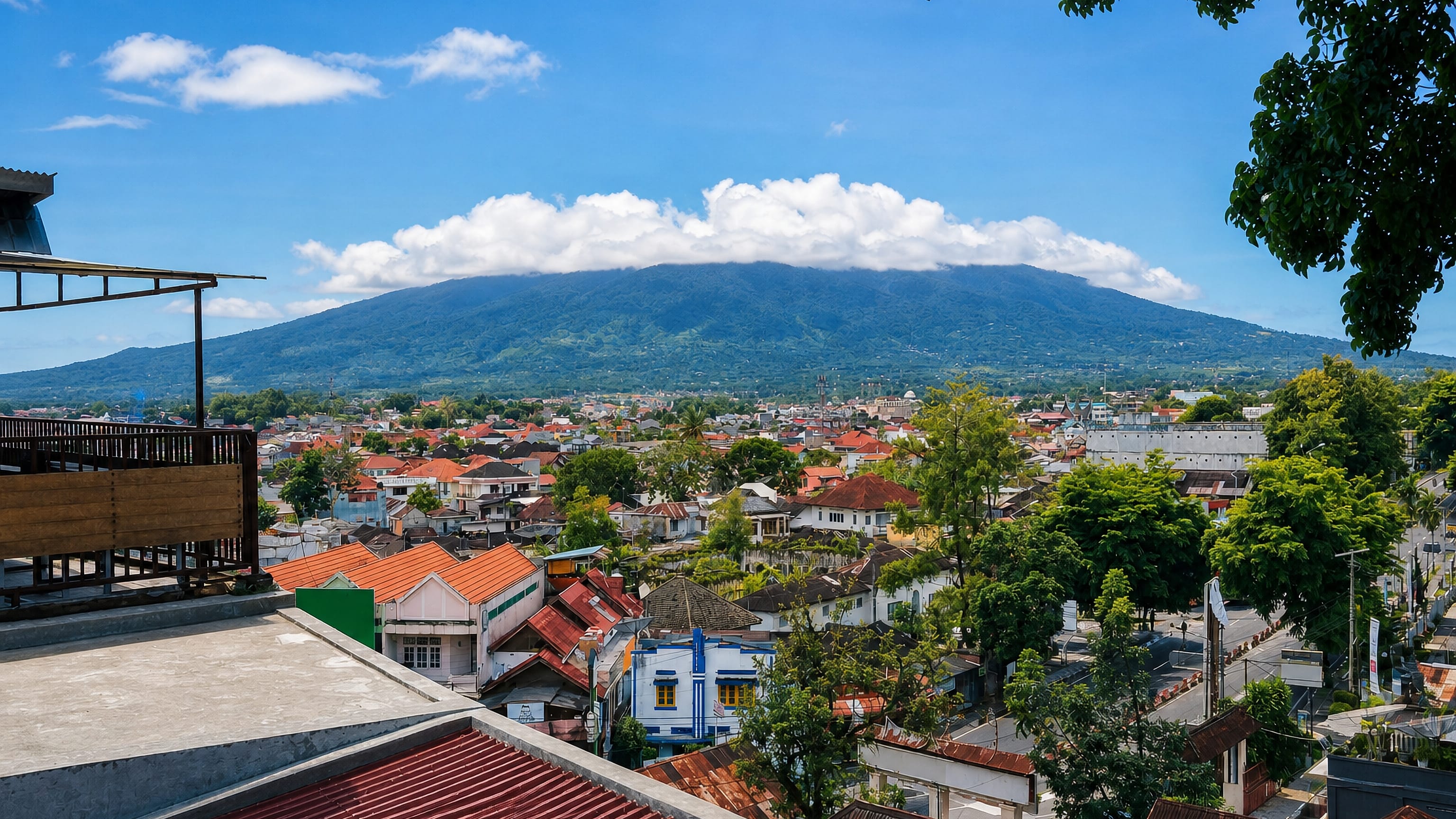 Rooftops of Bukittinggi, West Sumatra spreading below a cloud-capped volcano under a bright blue sky.