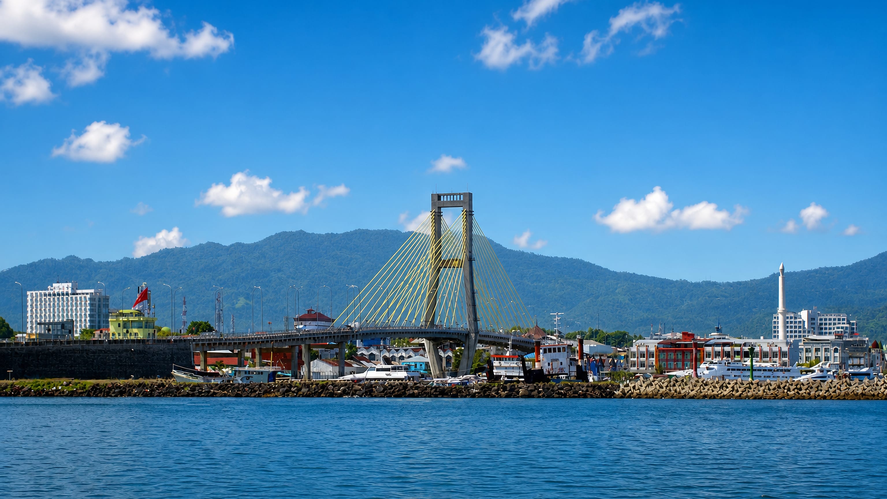 The cable-stayed Soekarno Bridge over Manado's harbour waterfront in Indonesia, with boats and green hills behind.