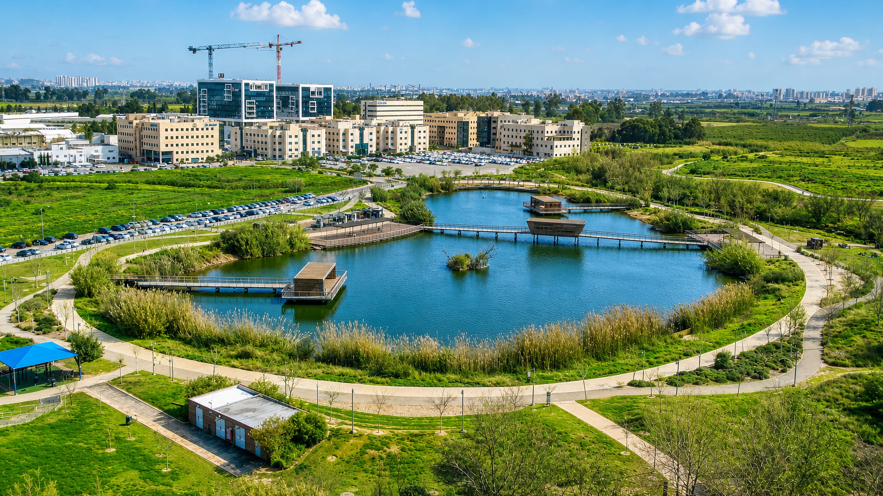 Aerial view of a lake park with boardwalks in Hod HaSharon, Israel, backed by office blocks and a distant skyline.