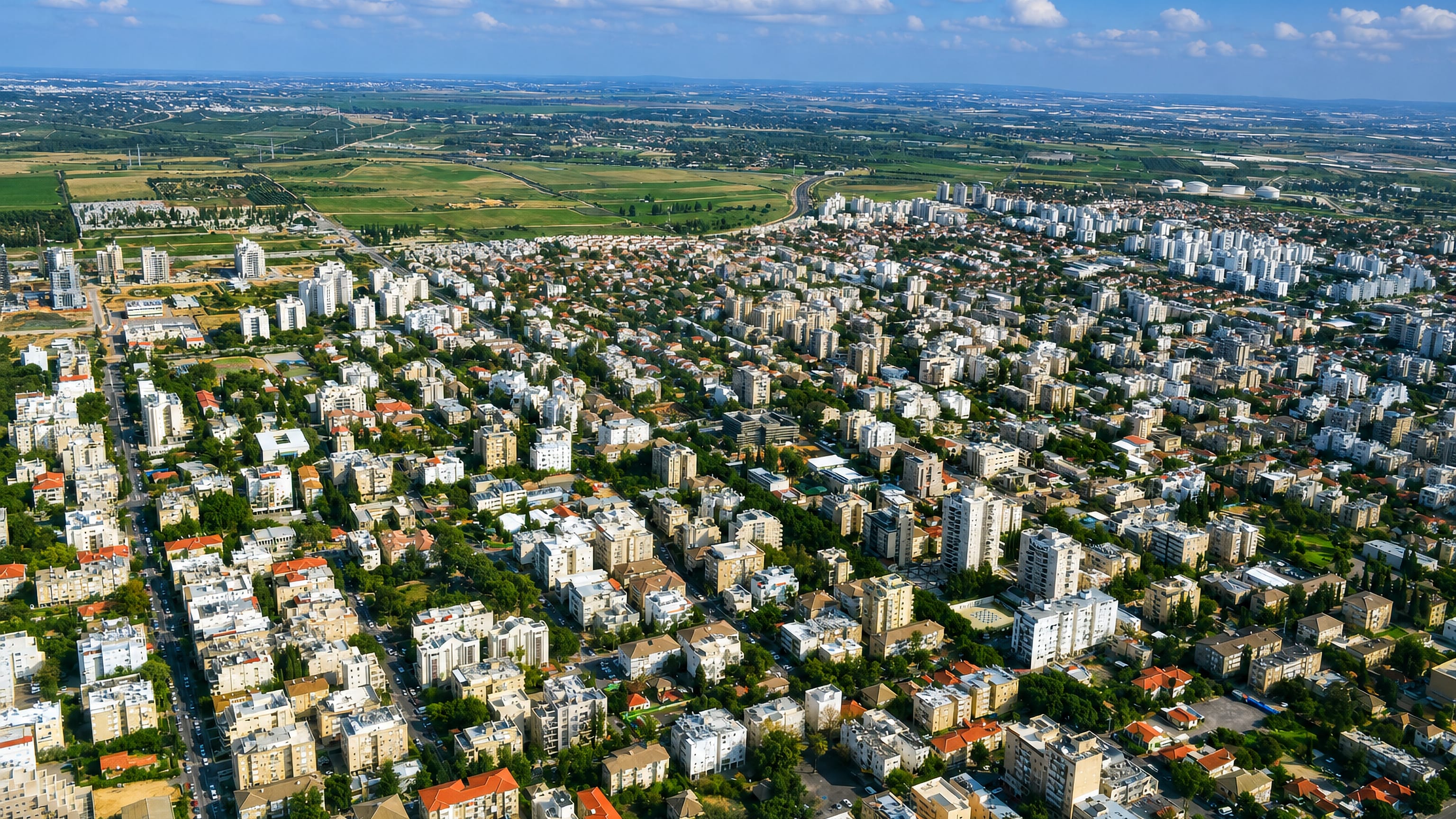 Aerial view over Rehovot, Israel, with dense white apartment blocks, red-tiled roofs and farmland on the horizon.