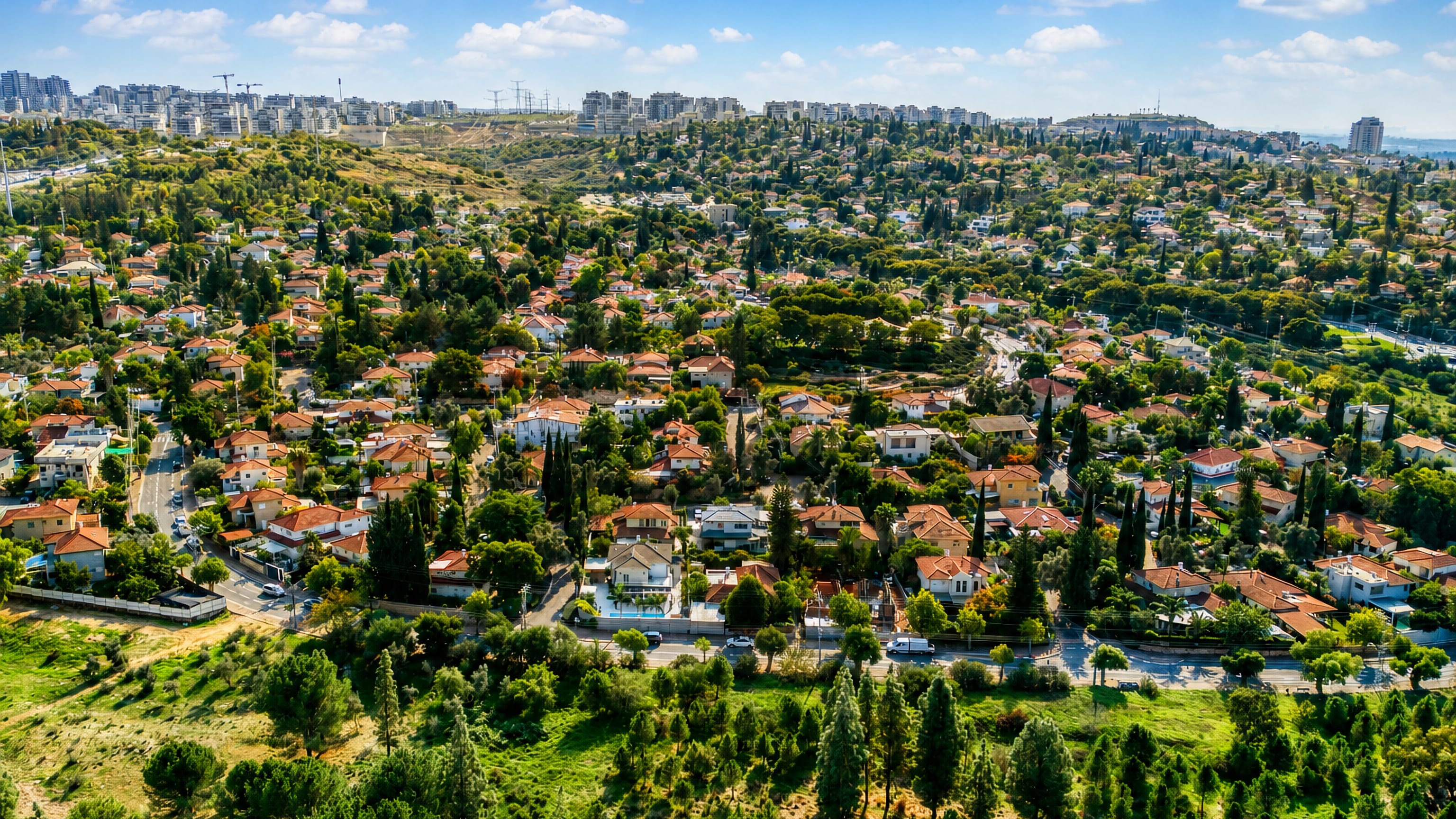 Aerial view of red-tiled villas and cypresses in Rosh Ha'Ayin, Israel, with apartment towers on the horizon.