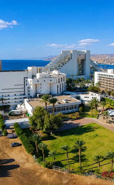Aerial view of Eilat's Red Sea resort hotels and palm-lined grounds with desert mountains beyond.