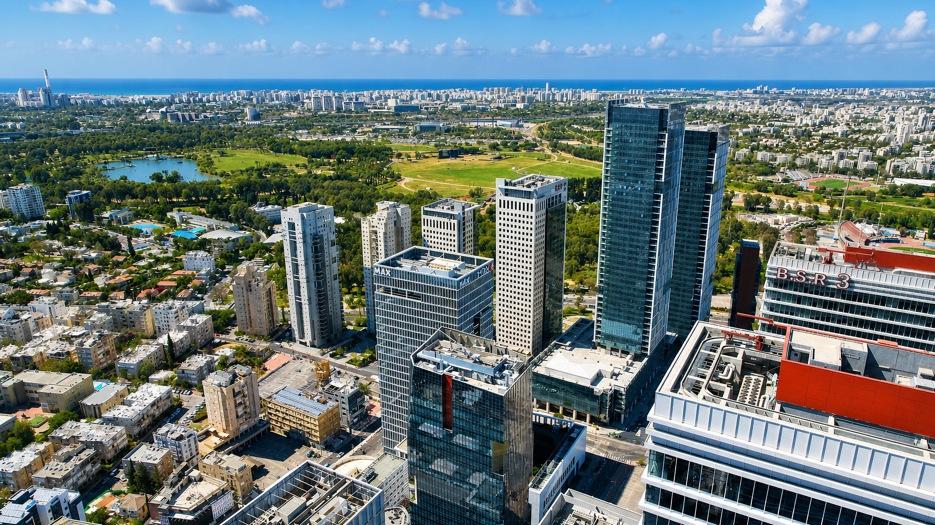 Aerial view of Bnei Brak's business district in Israel, glass office towers beside a green park and the sea beyond.