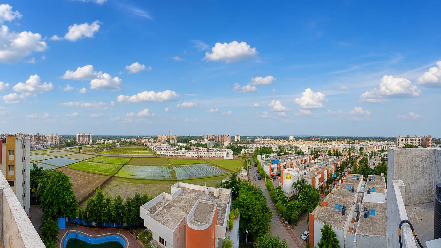 Rooftop panorama over Bhopal's suburban houses and green paddy fields under a wide blue sky, Madhya Pradesh.