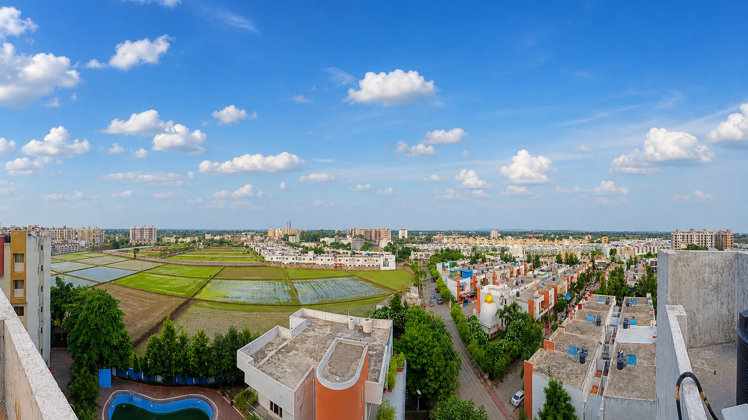 Rooftop panorama over Bhopal's suburban houses and green paddy fields under a wide blue sky, Madhya Pradesh.