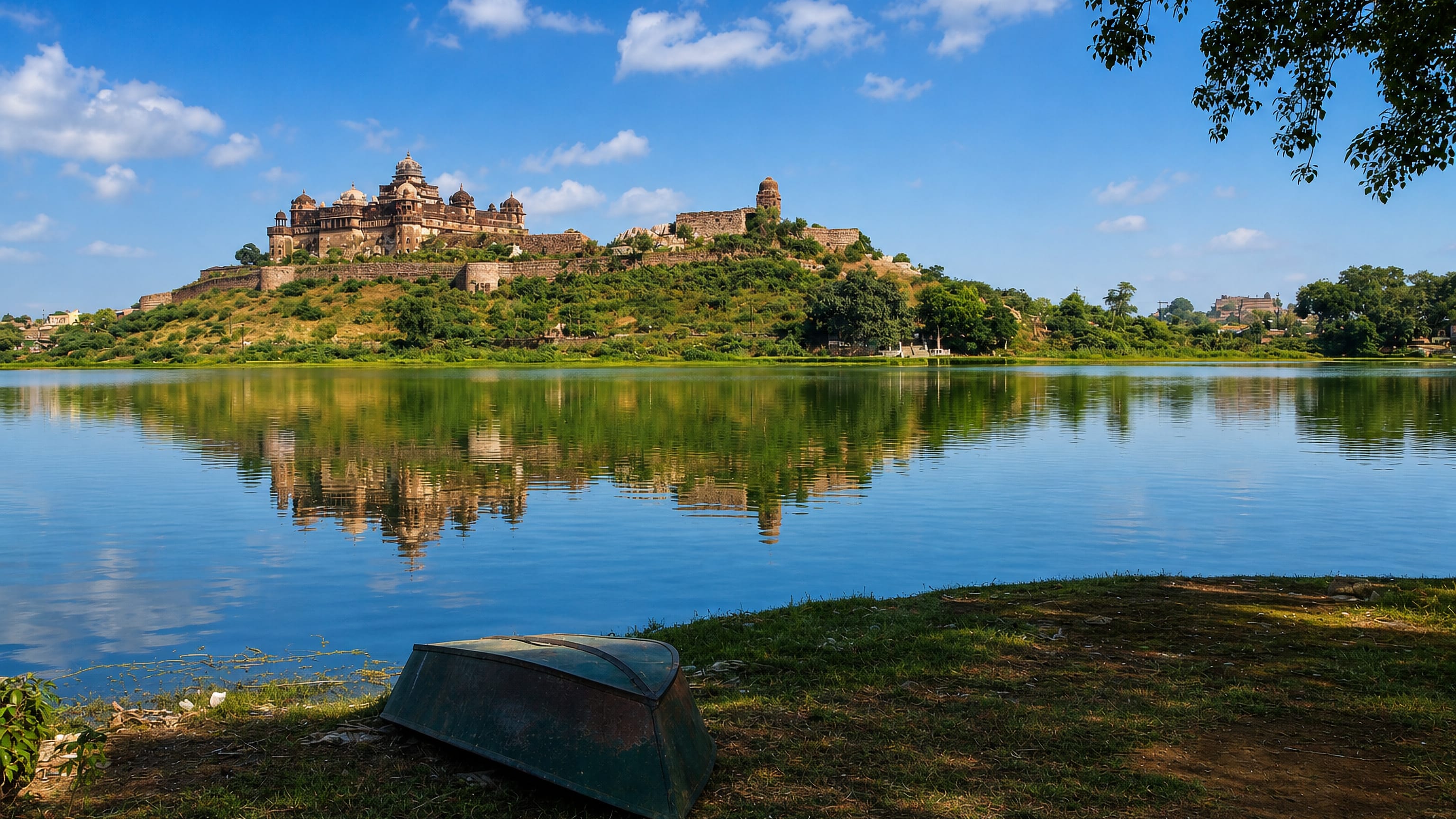 Hilltop palace and fort walls above a lake at Datia, Madhya Pradesh, with an upturned boat on the grassy shore.