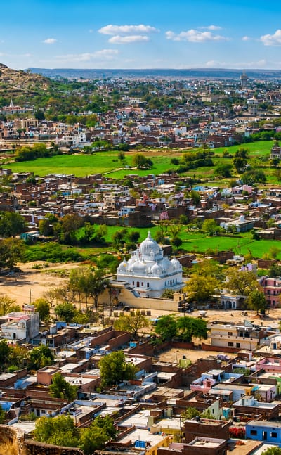 Aerial view over Gwalior's dense rooftops and a white domed shrine below a rocky hilltop fort, Madhya Pradesh.