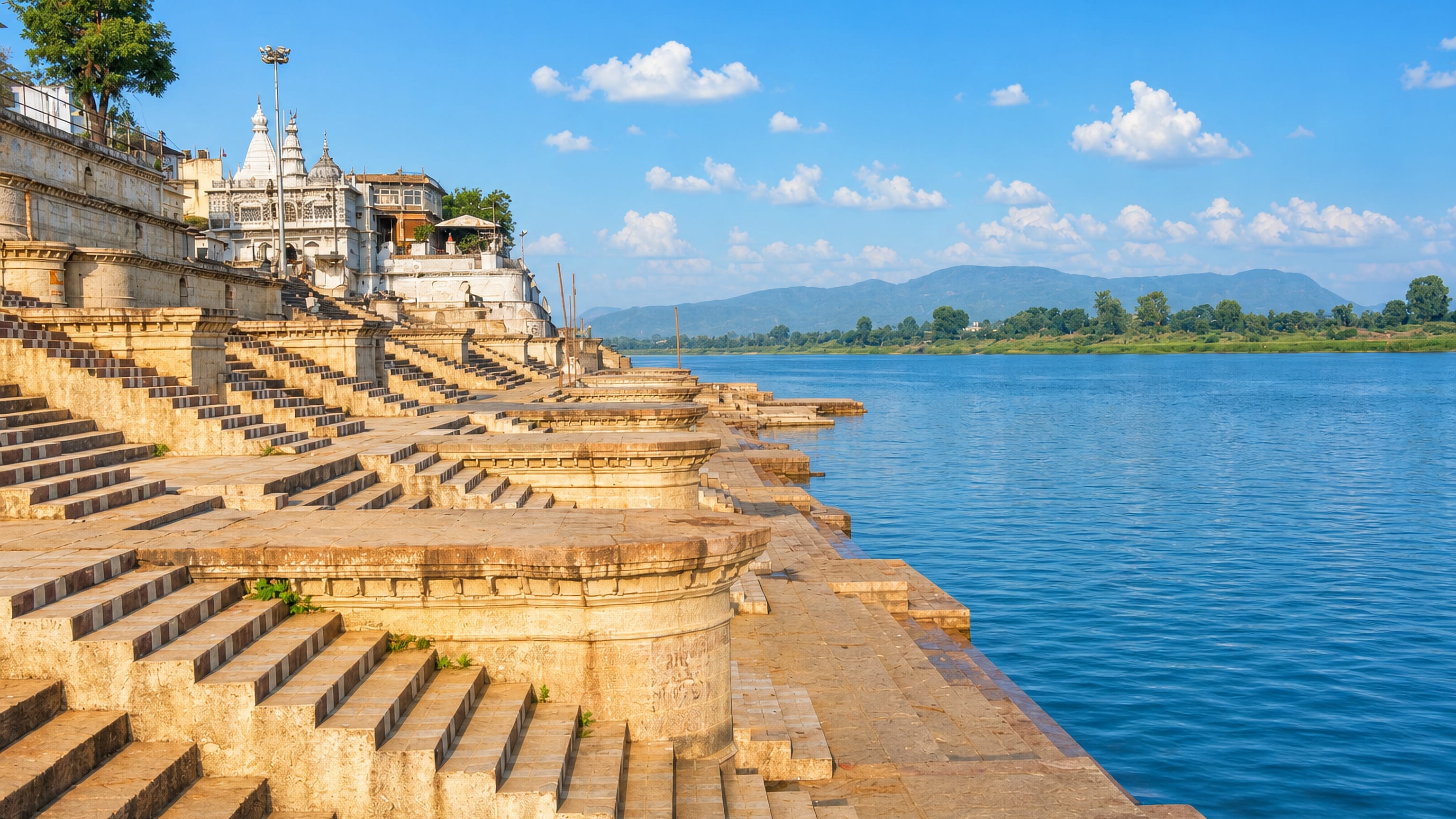 Stone ghat steps on the Narmada river at Narmadapuram, India, with a white temple and distant hills.