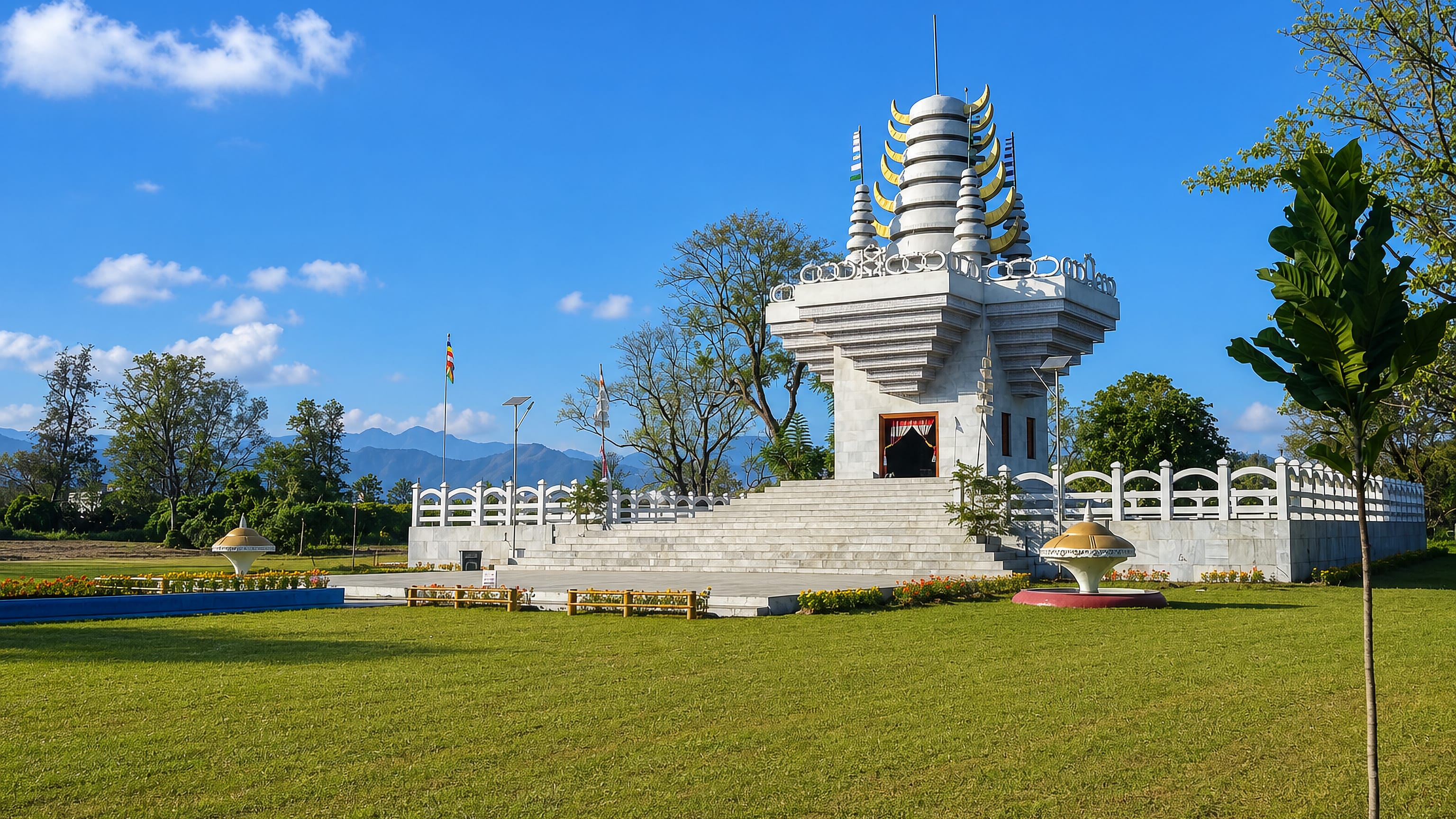 A white marble Manipuri-style memorial with tiered gold-tipped spires on a lawn in Imphal, hills behind.