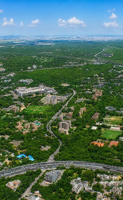 Aerial view over the leafy low-rise expanse and looping road interchanges of Delhi under a blue sky.