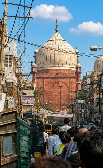 Crowded Old Delhi market lane of shop signs and wires beneath a Mughal domed mosque at Chawri Bazar, Delhi.
