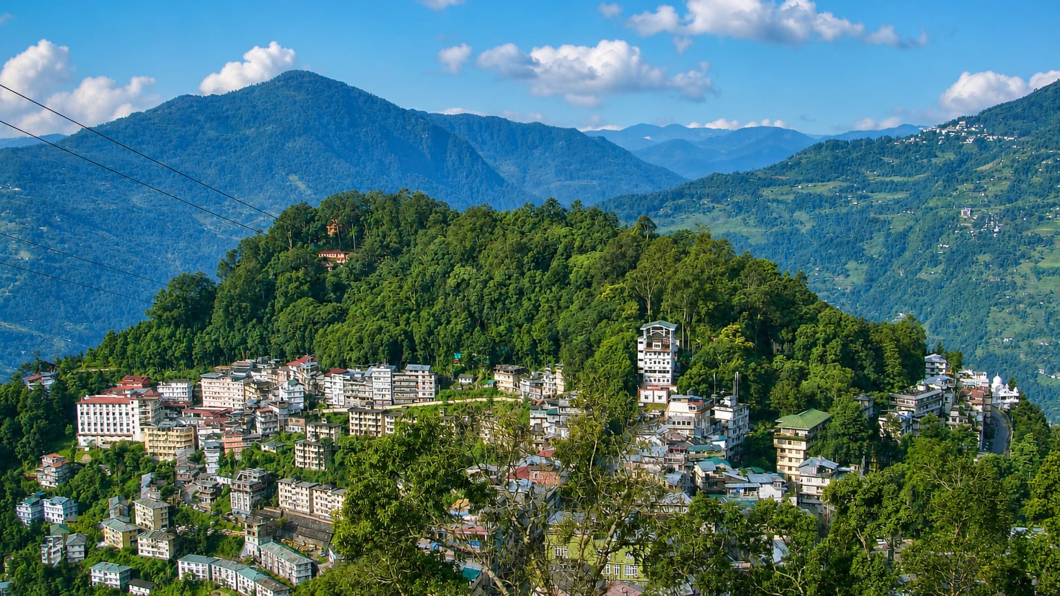 Gangtok's hillside houses on a forested ridge with a ropeway cable and Himalayan peaks beyond in Sikkim.