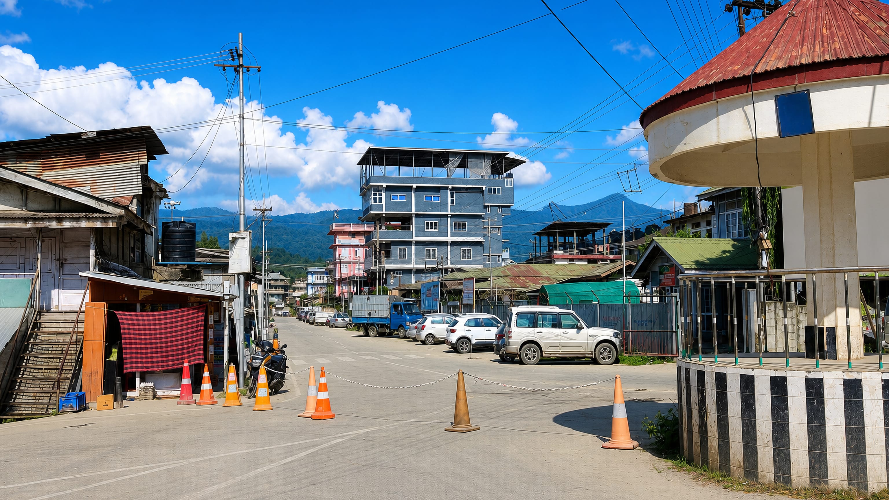 Quiet main street in Itanagar, Arunachal Pradesh, with parked SUVs, tin-roofed shops and forested hills beyond.