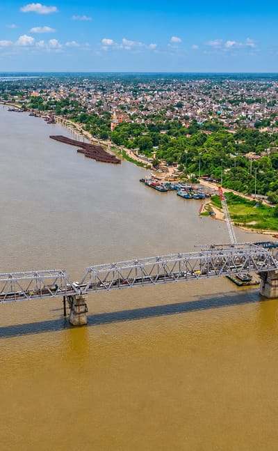 A long steel truss rail bridge spans the wide Ganges River beside Patna's low-rise cityscape under a blue sky.