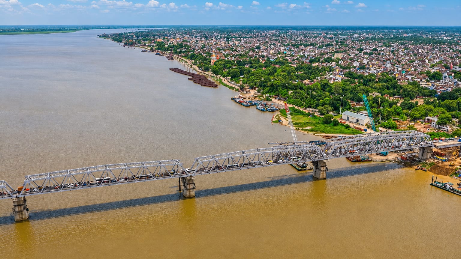 A long steel truss rail bridge spans the wide Ganges River beside Patna's low-rise cityscape under a blue sky.