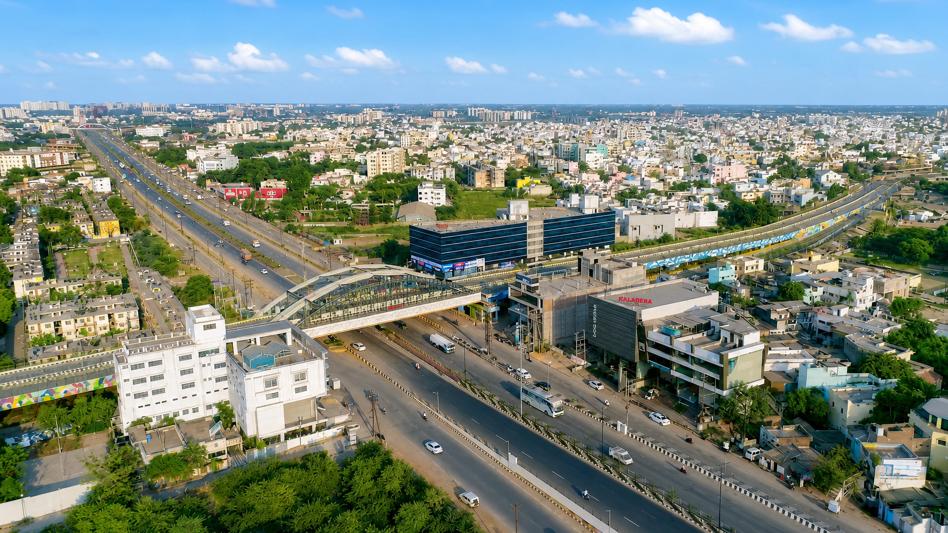 Aerial view over Raipur, India, showing a wide multi-lane boulevard, pedestrian skywalk and low-rise city sprawl.