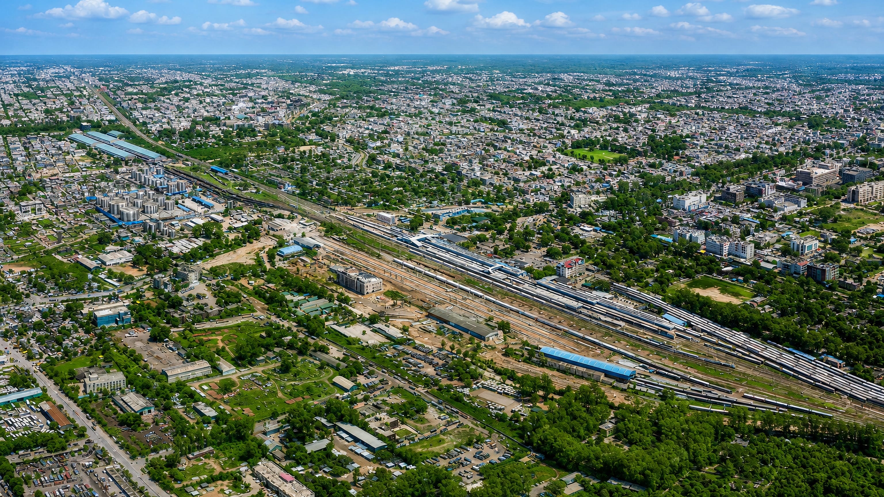 Aerial view over Jammu, India, showing dense low-rise neighbourhoods flanking a wide railway yard under a bright sky.
