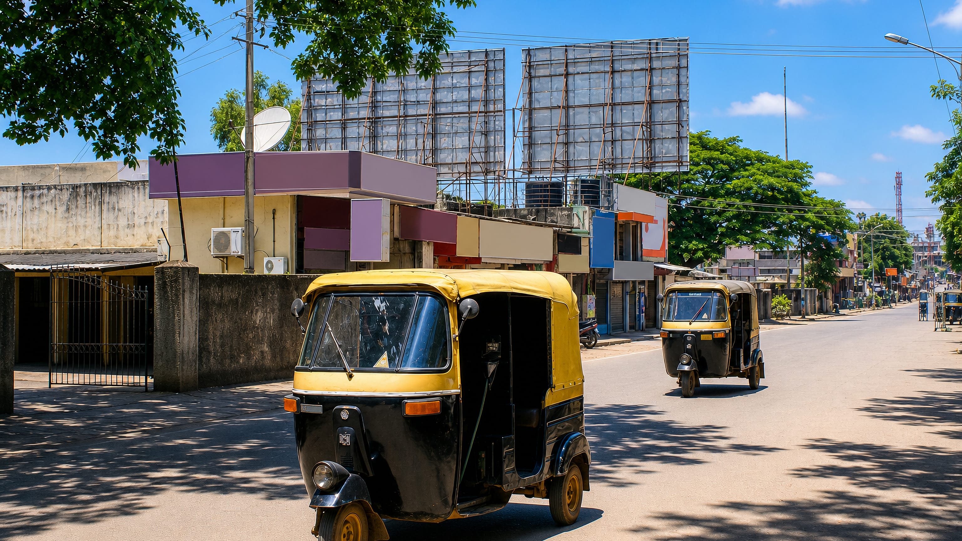Auto-rickshaws on a quiet shopfront street in Hassan, Karnataka, beneath empty billboard frames.