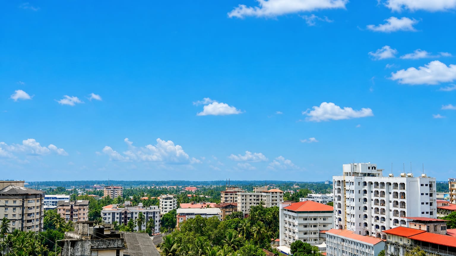 Red-tiled roofs and coconut palms among Mangaluru's white mid-rise buildings under a blue sky.