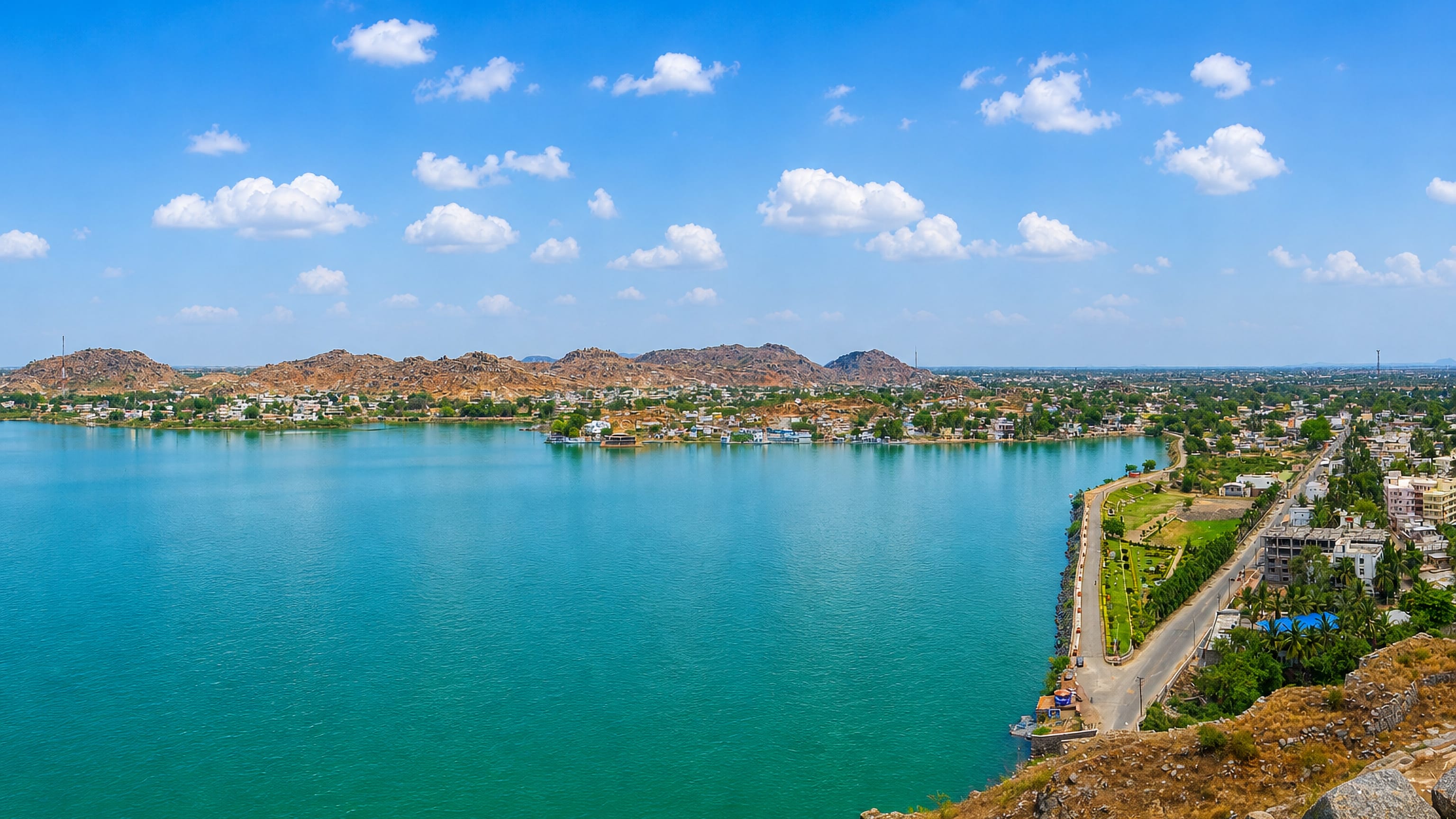 Turquoise reservoir ringed by rocky Deccan hills, with the edge of Raichur town along the far shore.