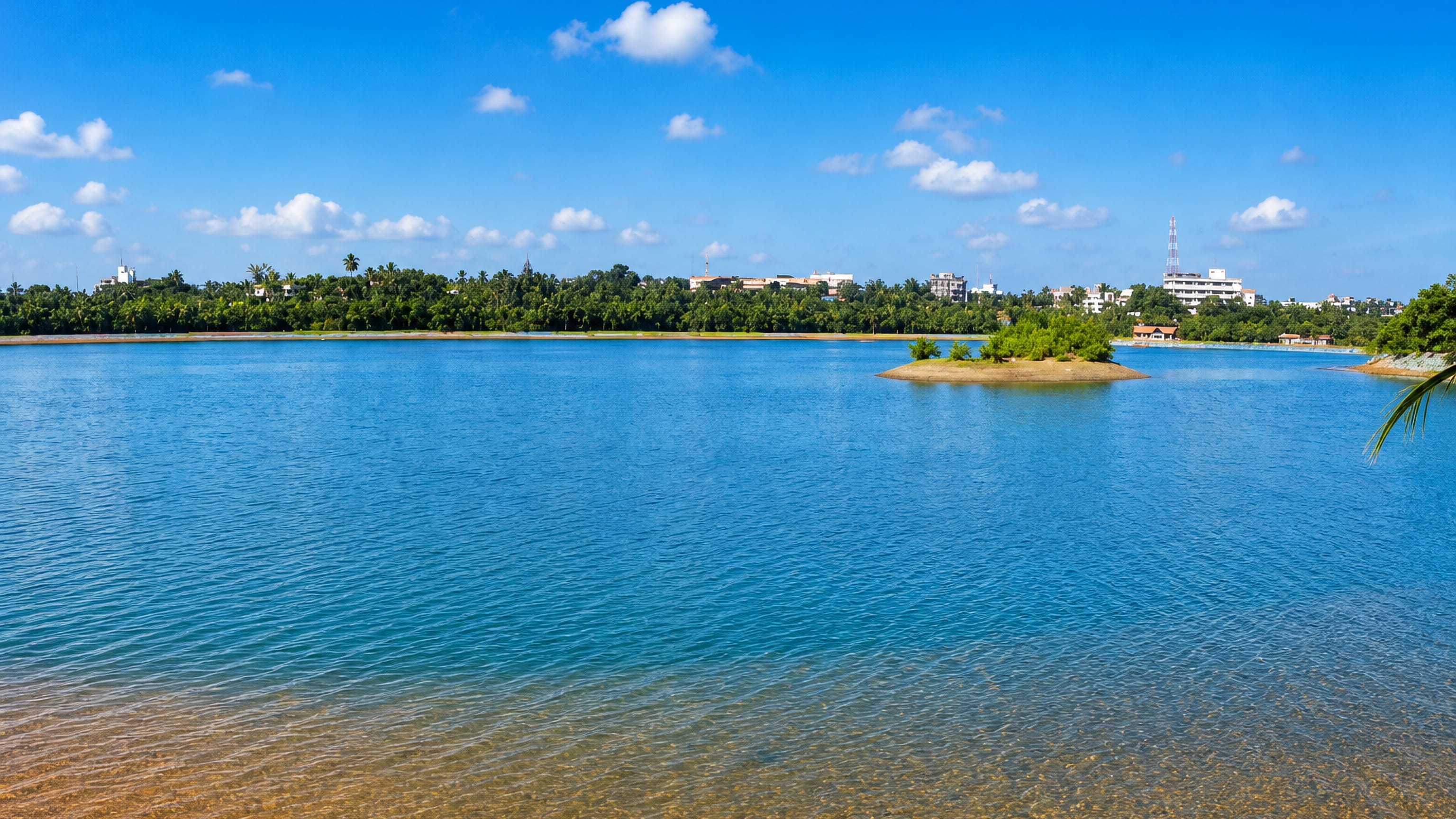 Calm blue lake with a small island fringed by palms below the low skyline of Manipal in coastal Karnataka