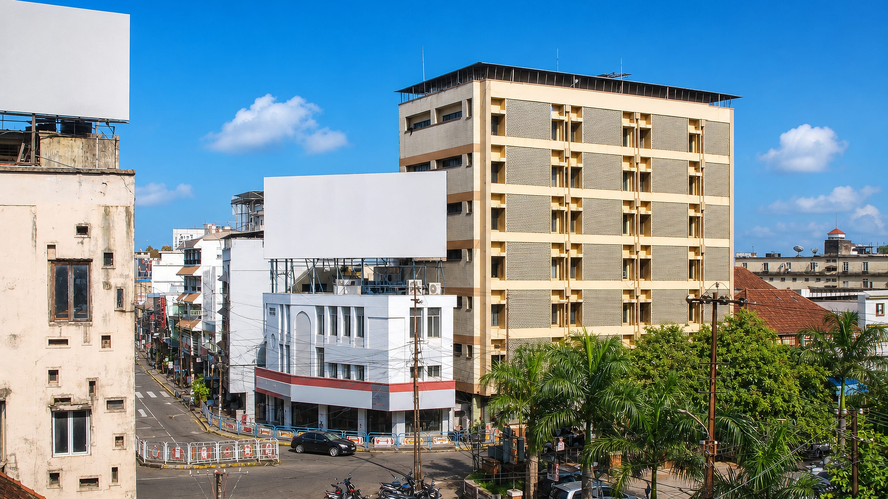Street corner in Kottayam, Kerala, with a mid-rise apartment block, billboards and parked motorbikes.