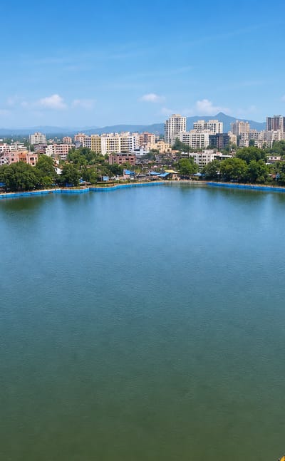 A large lake ringed by mid-rise buildings and green hills at Kalyan under a bright blue sky.