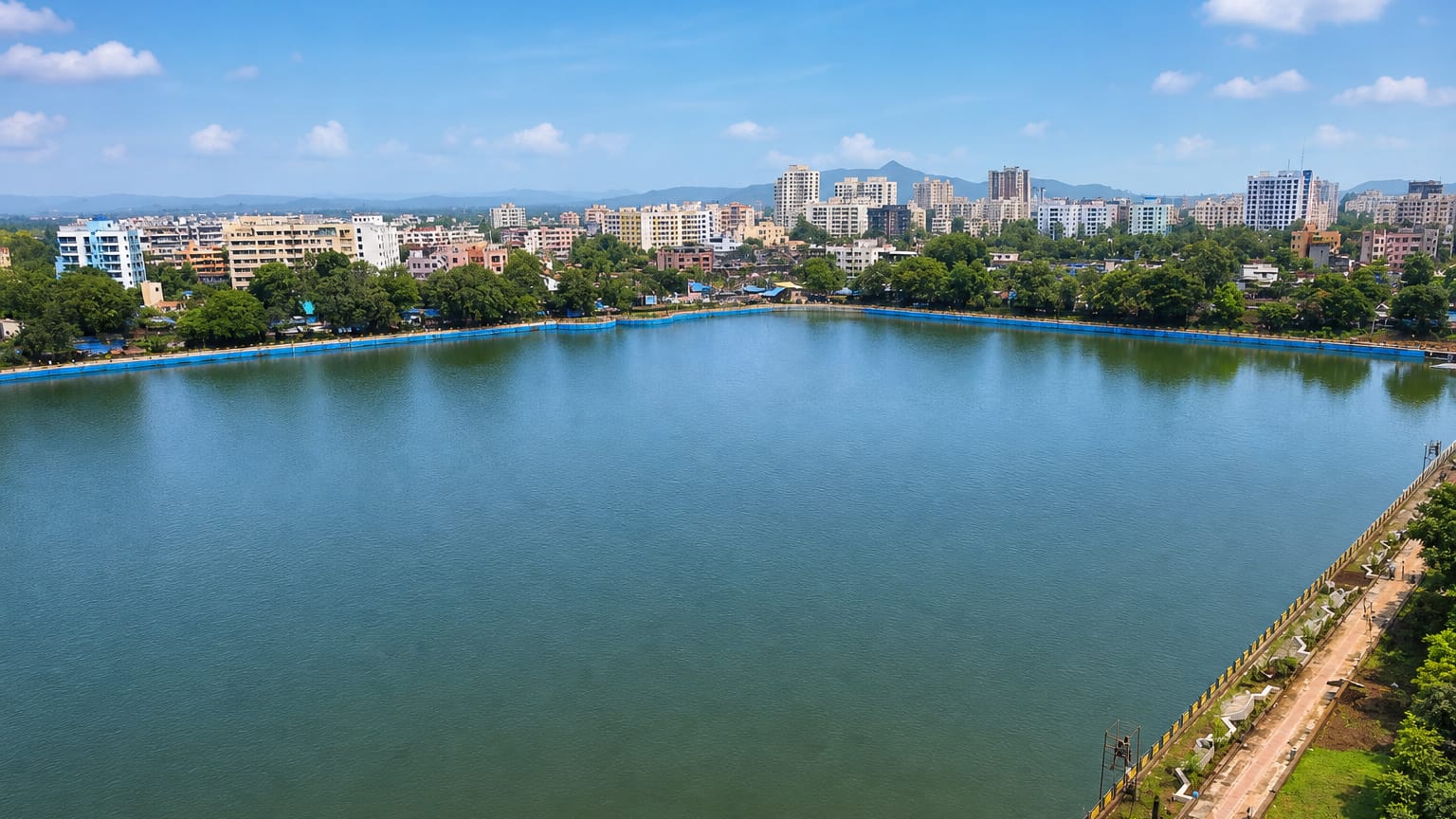 A large lake ringed by mid-rise buildings and green hills at Kalyan under a bright blue sky.